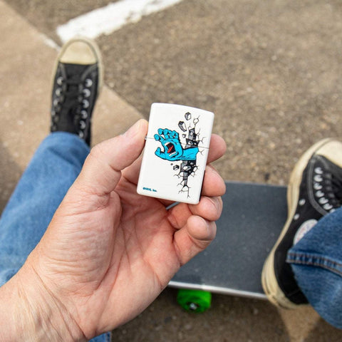 Lifestyle image of Santa Cruz Screaming Hand White Matte Windproof Lighter closed in hand, with a skateboard in the background