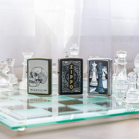 Lifestyle image of Zippo Filigree Design Black Matte Windproof Lighter standing on a glass chess board with a Skull lighter, a Chess Themed lighter, and glass chess pieces