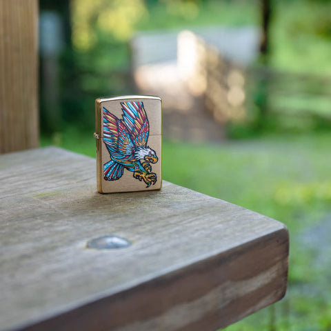 Lifestyle image of Tattoo Eagle Design Brushed Brass Windproof Lighter standing on a railing with trees and a bridge in the background.