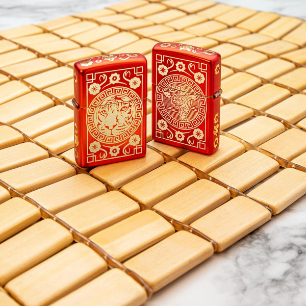 Lifestyle image of two Tiger Design Metallic Red Windproof Lighters, one showing the front and the other showing the back of the lighter. The lighters are standing on a mat.